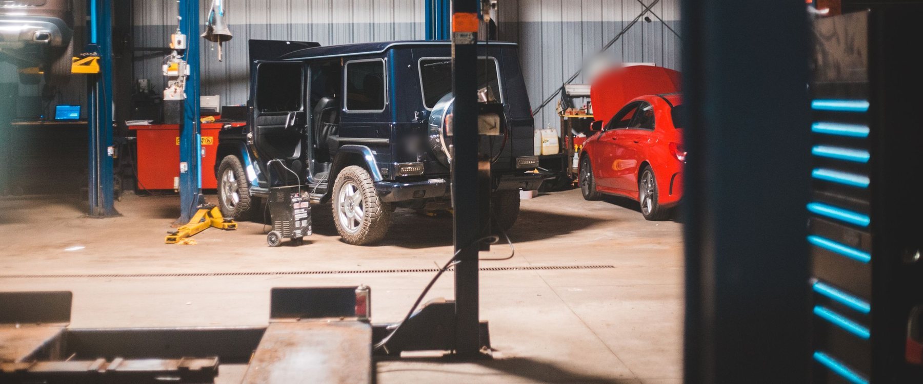 Oficina ampla com elevadores azuis, um utilitário escuro de portas abertas e um carro vermelho de capô levantado ao fundo
