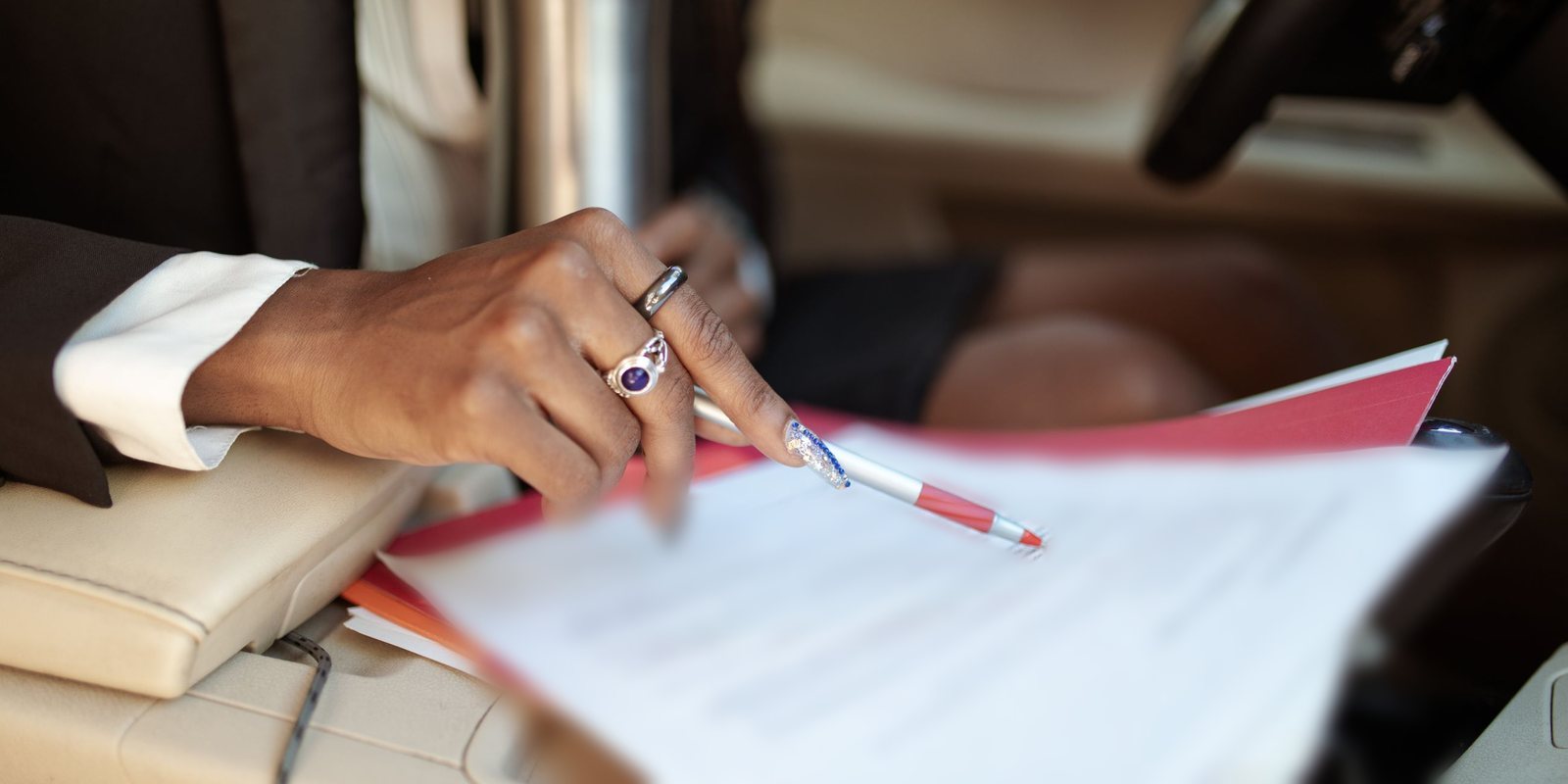 Mão com caneta vermelha sobre folhas impressas apoiadas no banco de um carro