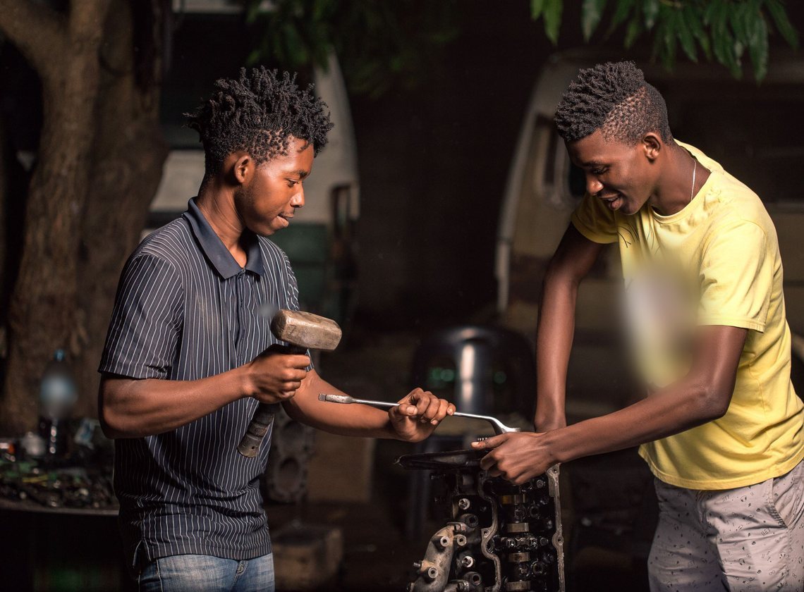 Dois homens trabalham juntos num cabeçote de motor, um com martelo e o outro com chave de boca, à noite, sob uma árvore